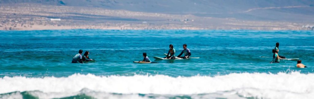 Clases de surf en la Playa de Famara, Lanzarote, con surfistas en el océano y montañas al fondo