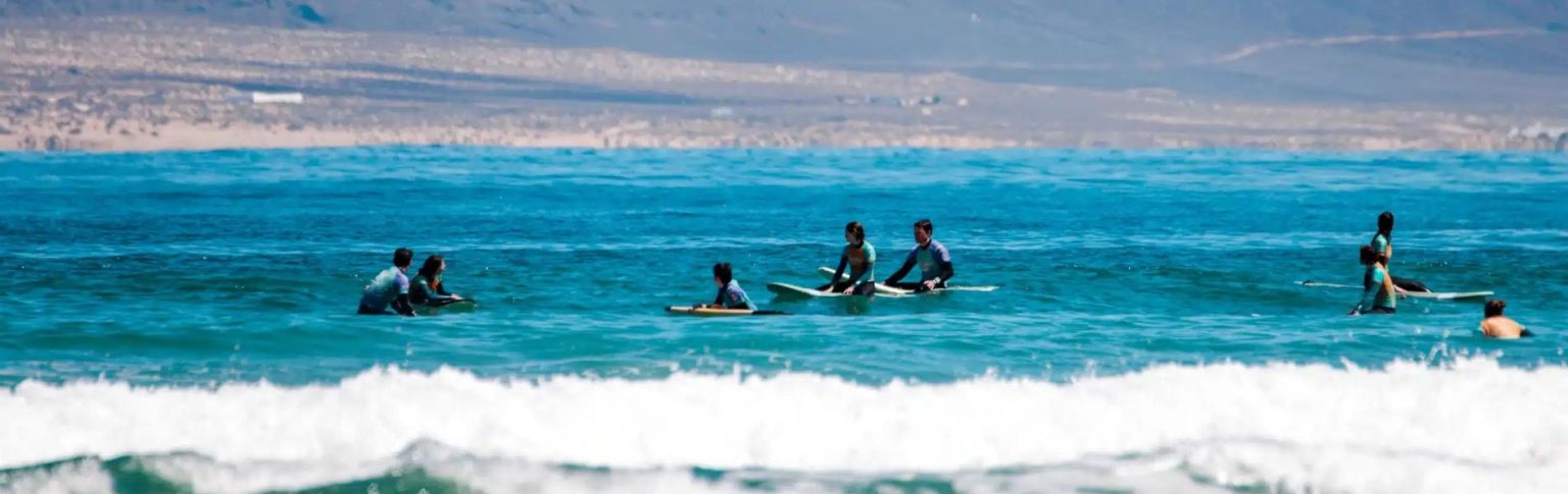 Clases de surf en la Playa de Famara, Lanzarote, con surfistas en el océano y montañas al fondo