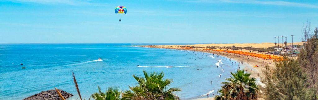 Playa del Inglés en Gran Canaria con dunas, parasailing, sombrillas naranjas y aguas turquesas en día soleado