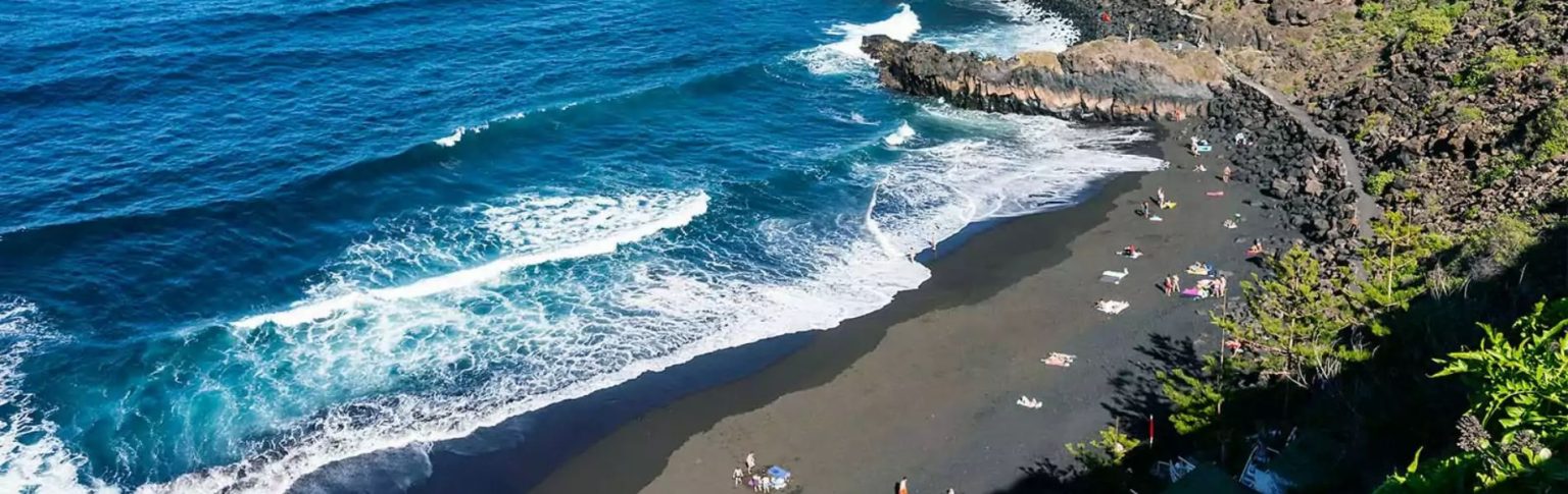 Playa El Bollullo con arena volcánica negra y aguas turquesas en Tenerife, vista aérea con bañistas