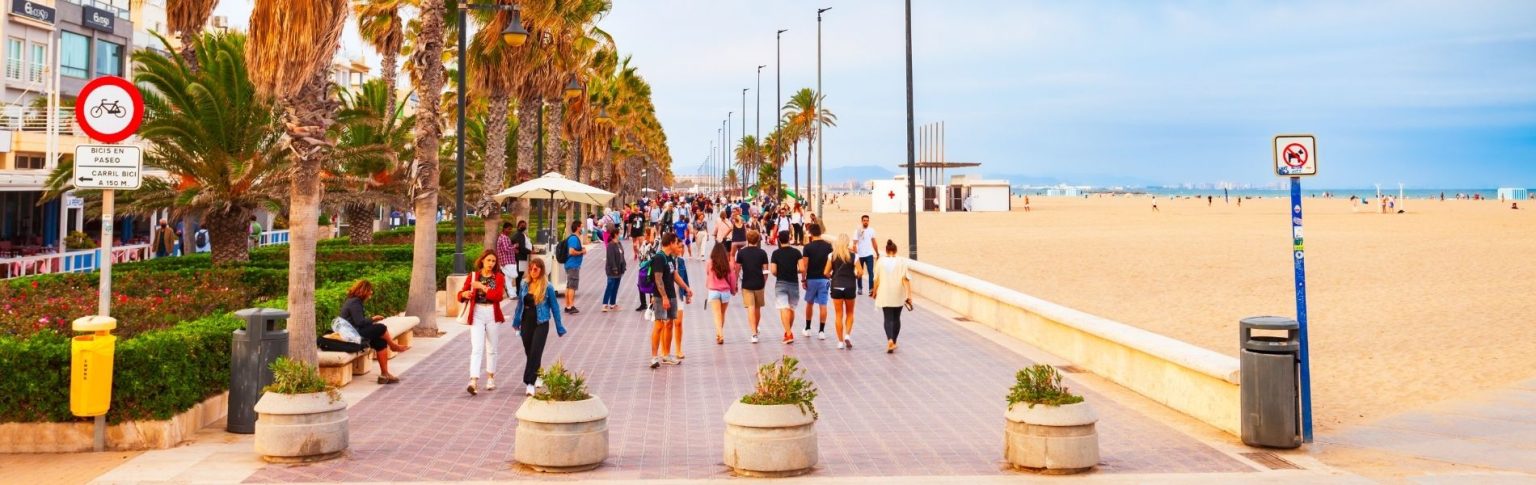 Paseo marítimo de Valencia con palmeras, turistas paseando y la playa de arena dorada al fondo en un día soleado