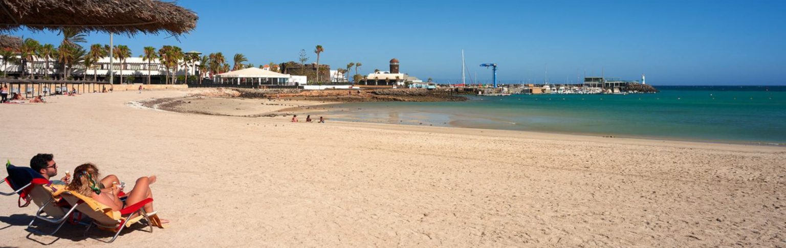 Playa del Castillo en Fuerteventura con arena dorada, aguas turquesas y pareja tomando el sol bajo palmeras