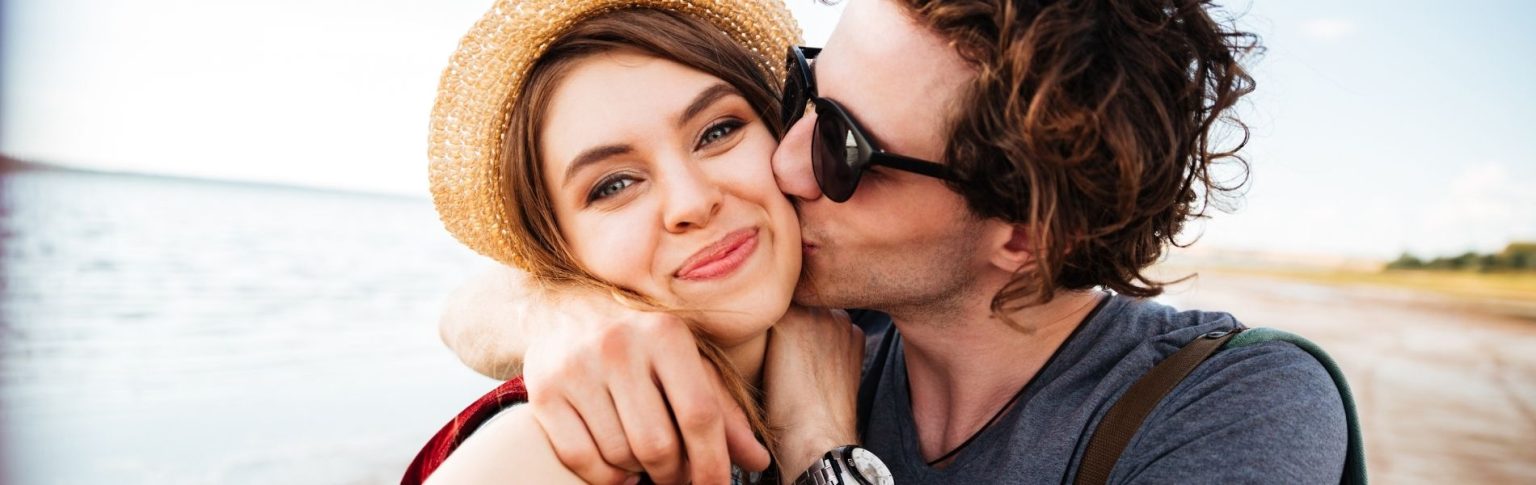 Pareja joven enamorada junto al mar, él dándole un beso en la mejilla a ella con sombrero de paja