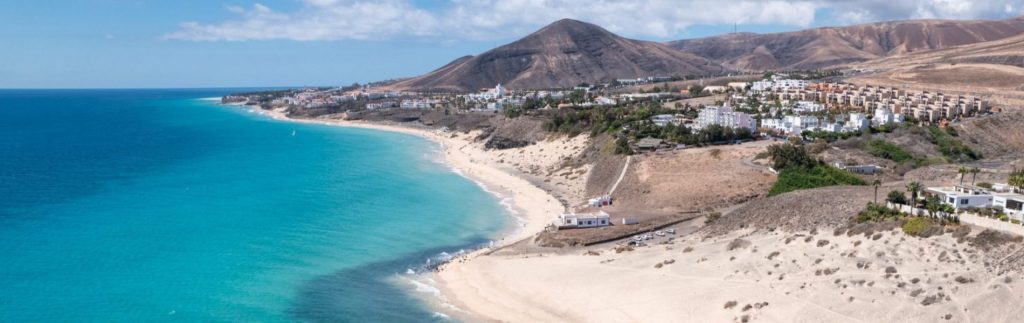 Vista aérea de la Playa de Esquinzo en Fuerteventura con aguas turquesas, arena blanca y montañas volcánicas al fondo